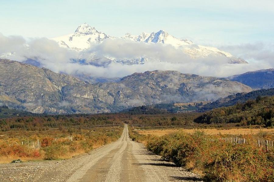 Carretera Austral, Chile - Dirt road leading straight to snowy mountains in the distance. Blue sky. Low clouds veiling the middle elevations of the mountains.